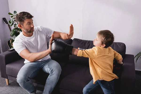 Cute boy in boxing gloves fighting with father sitting on sofa — Stock Photo