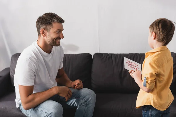 Happy father sitting on sofa near adorable son holding happy fathers day greeting card — Stock Photo