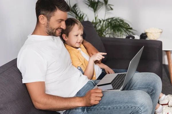 Lächelnder Junge zeigt mit dem Finger auf Laptop, während er neben lächelndem Vater sitzt — Stockfoto