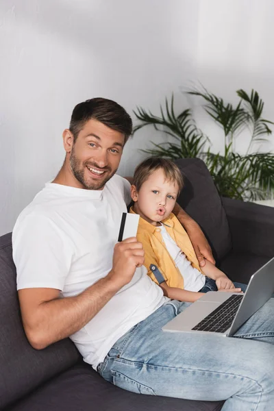 Усміхнений батько показує кредитну картку, сидячи з ноутбуком біля похмурого сина — стокове фото