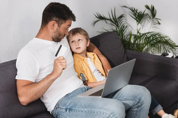 Vater hält Kreditkarte in der Hand und schaut den niedlichen Sohn an, während er mit Laptop auf dem Sofa sitzt — Stockfoto