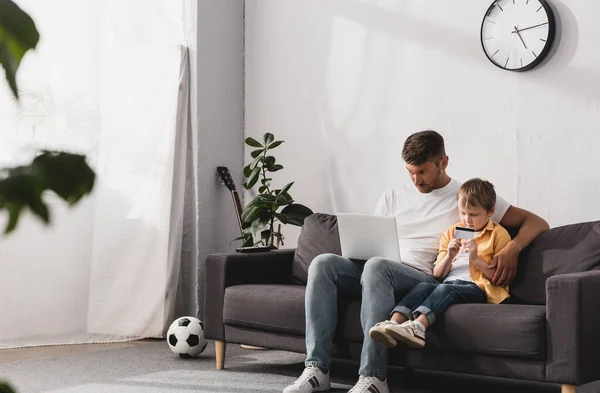 Selective focus of father using laptop and his son holding credit card while sitting on sofa at home — Stock Photo