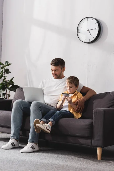 Hombre guapo usando el ordenador portátil mientras que su hijo lindo celebración de la tarjeta de crédito - foto de stock