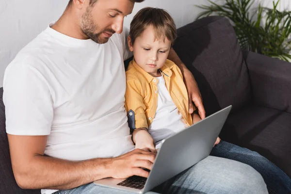 Padre e hijo atentos usando el ordenador portátil mientras está sentado en el sofá en casa - foto de stock