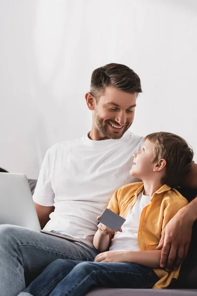 Счастливый отец с ноутбуком глядя на улыбающийся сын держит кредитную карту — стоковое фото
