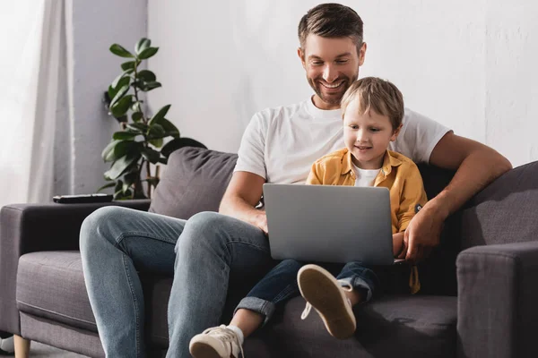 Feliz padre e hijo sentado en el sofá y el uso de la computadora portátil juntos - foto de stock