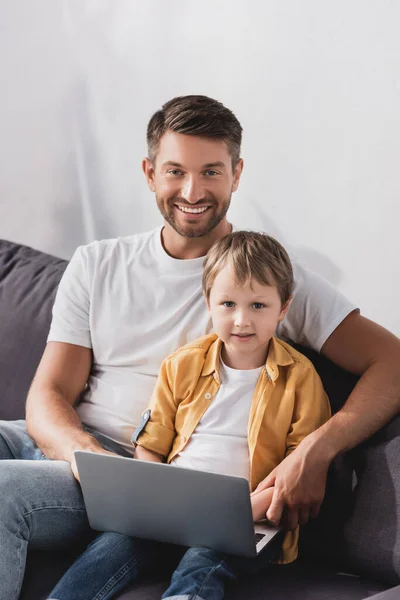 Feliz padre e hijo sonriendo a la cámara mientras usa el ordenador portátil en casa - foto de stock