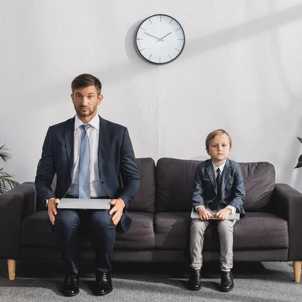 Serious businessman and his son in formal wear sitting with laptops on sofa at home and looking at camera — Stock Photo