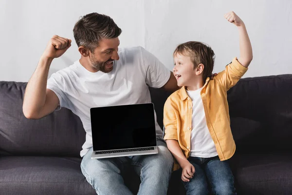 Padre e hijo emocionados mostrando gesto ganador mientras se sienta con el ordenador portátil con pantalla en blanco - foto de stock