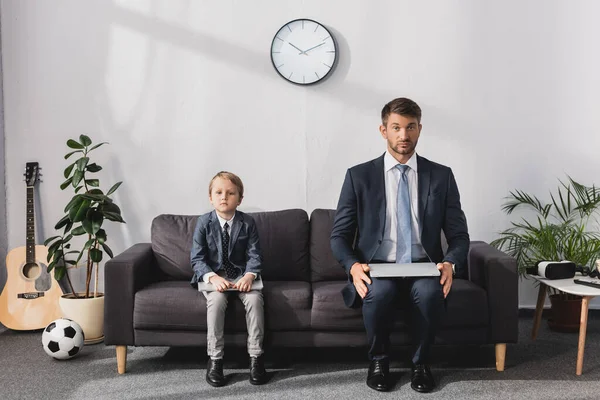 Hombre de negocios serio y su hijo en ropa formal sentado con computadoras portátiles en el sofá en casa y mirando a la cámara - foto de stock
