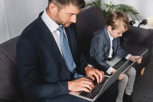 Hombre de negocios serio y su hijo en ropa formal usando computadoras portátiles en casa - foto de stock