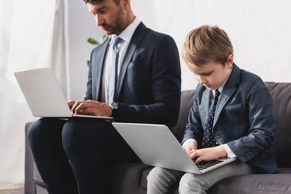 Hombre de negocios guapo y su hijo en ropa formal usando computadoras portátiles en casa - foto de stock