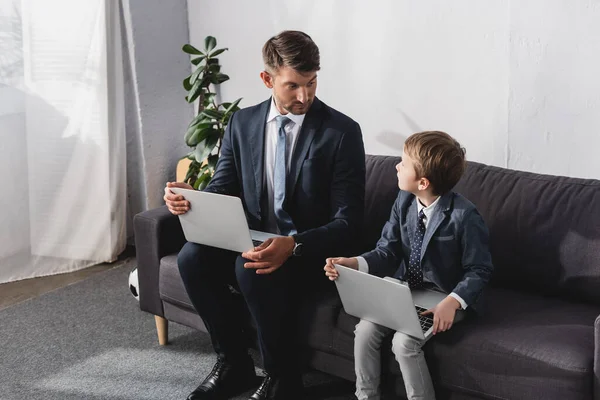 Hombre de negocios serio con su hijo en ropa formal sentado en el sofá con computadoras portátiles - foto de stock