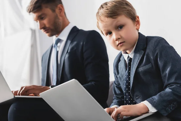 Adorable, chico serio y su padre en ropa formal usando computadoras portátiles en casa - foto de stock