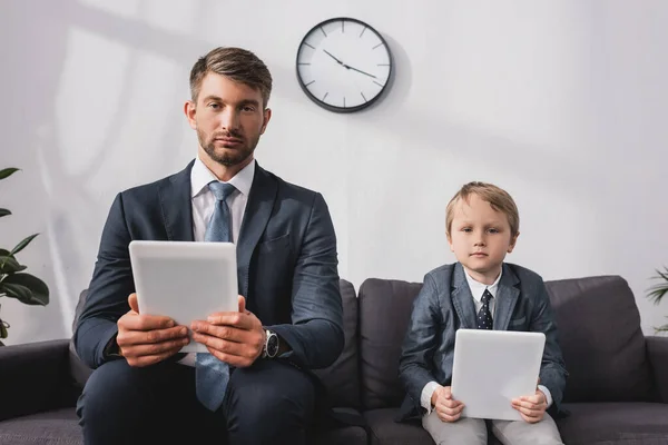 Hombre de negocios serio y su hijo en ropa formal sosteniendo tabletas digitales mientras está sentado en el sofá en casa - foto de stock