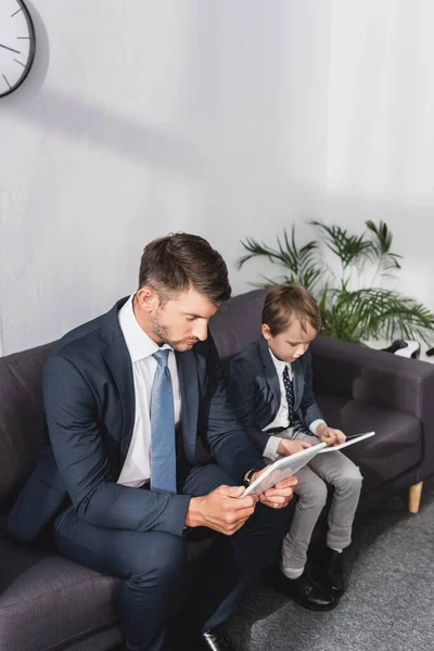Ориентированный бизнесмен и его сын в формальной одежде с помощью цифровых таблеток, сидя на диване дома — стоковое фото