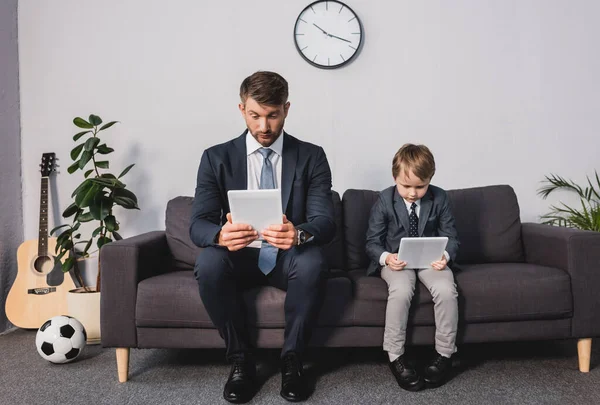 Homem de negócios concentrado e seu filho em desgaste formal usando tablets digitais enquanto sentado no sofá em casa — Fotografia de Stock