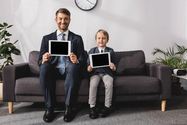 Homem de negócios feliz e filho no desgaste formal mostrando tablets digitais com tela em branco e olhando para a câmera — Fotografia de Stock