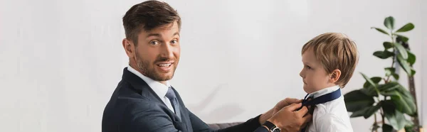 Panoramic crop of businessman smiling at camera while putting tie on adorable son at home — Stock Photo