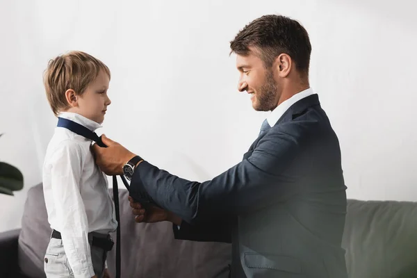 Side view of businessman in formal wear putting tie on adorable son at home — Stock Photo