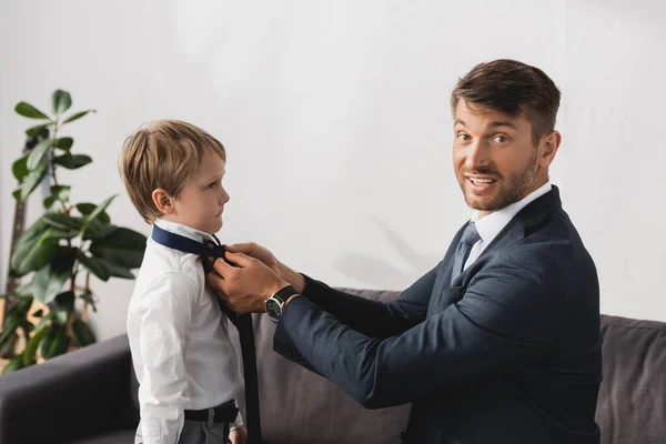 Lächelnder Geschäftsmann in offizieller Kleidung, der in die Kamera schaut, während er zu Hause seinen entzückenden Sohn mit Krawatte anzieht — Stockfoto