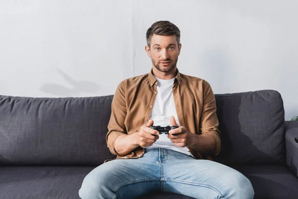 KYIV, UKRAINE - 9 JUIN 2020 : homme concentré assis sur le canapé et jouant à un jeu vidéo avec joystick — Photo de stock