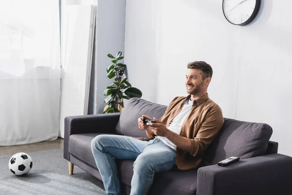 KYIV, UCRANIA - 9 de junio de 2020: hombre alegre jugando videojuegos con joystick mientras está sentado en el sofá cerca de la pelota de fútbol - foto de stock