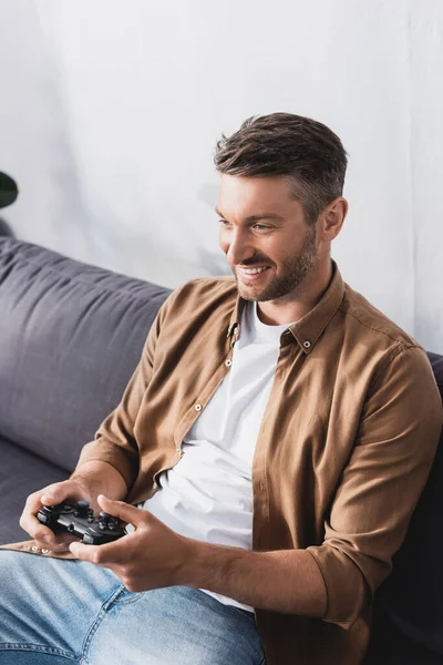KYIV, UCRANIA - 9 de junio de 2020: hombre feliz y guapo sentado en el sofá y jugando videojuegos con joystick - foto de stock