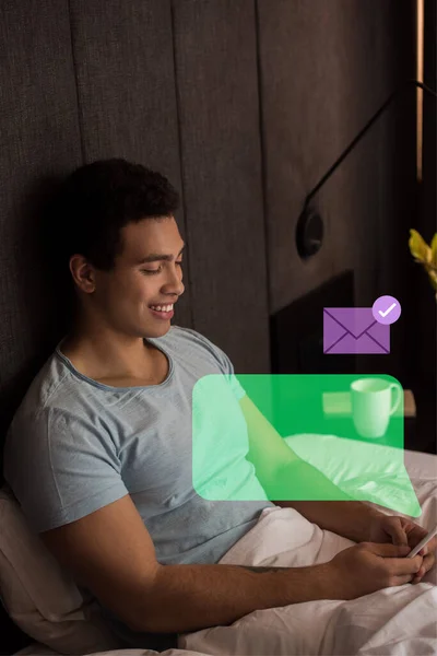 Glücklich gemischter Rennmann mit Smartphone in der Nähe von virtueller Sprechblase und Briefumschlag im Schlafzimmer — Stockfoto