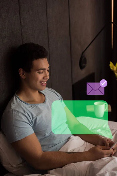 Happy mixed race man using smartphone near virtual speech bubble and envelope in bedroom — Stock Photo