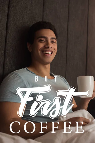 Smiling mixed race man holding cup of coffee near but first coffee lettering in bedroom — Stock Photo