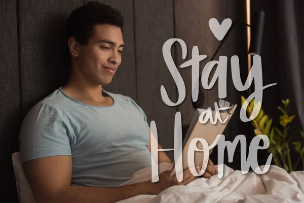Smiling mixed race man reading book near stay at home lettering in bedroom — Stock Photo