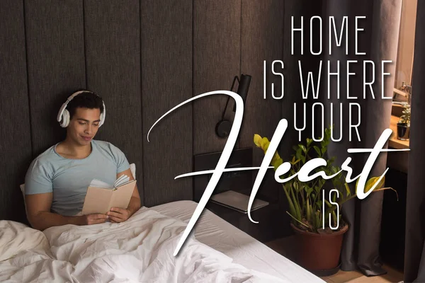 Handsome mixed race man in headphones listening audio book and holding book near home is where your heart is lettering in bedroom — Stock Photo