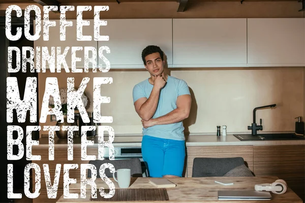 Pensoso uomo razza mista in piedi vicino a tazza di caffè, libro, cuffie, laptop e bevitori di caffè fanno meglio lettering amanti — Foto stock