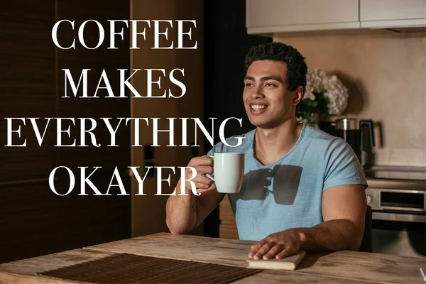 Hombre de raza mixta feliz sosteniendo la taza de café cerca de libro y café hace que todo letras okayer - foto de stock