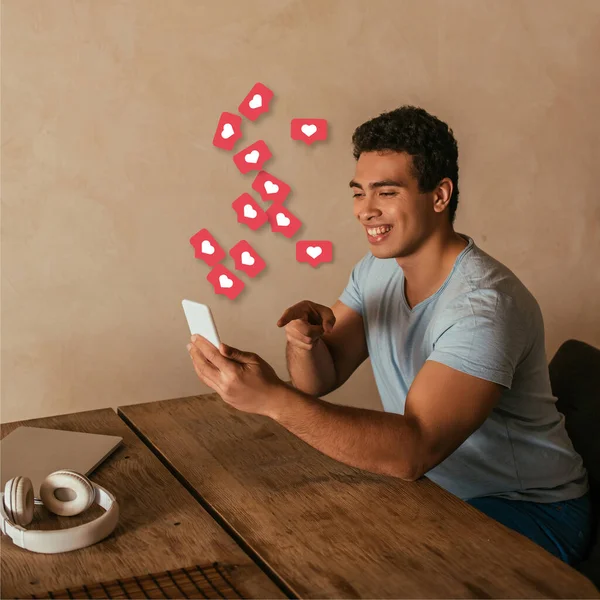 Happy mixed race man pointing with finger at smartphone, laptop, headphones and virtual hearts illustration — Stock Photo