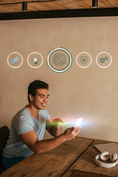 Hombre de raza mixta feliz apuntando con el dedo al teléfono inteligente, portátil, auriculares, huella digital e iconos virtuales ilustración - foto de stock