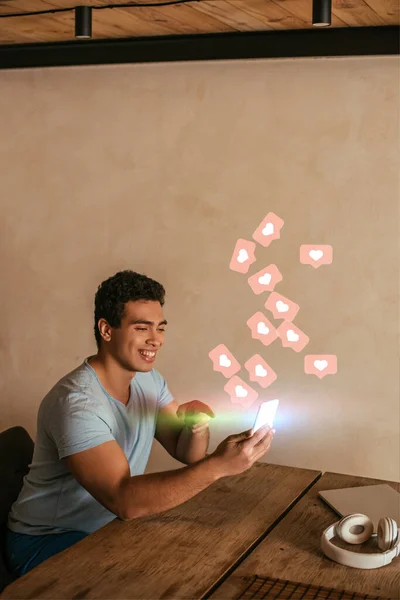 Счастливый смешанный человек, показывающий пальцем на смартфон, ноутбук, наушники и виртуальные сердца в качестве иллюстрации — стоковое фото