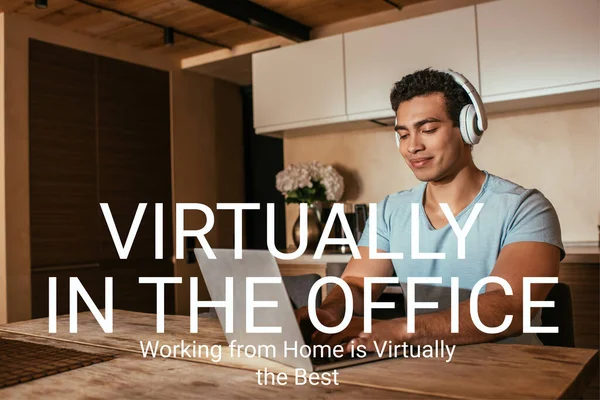 Happy mixed race freelancer listening music in headphones and using laptop near virtually in the office, working from home is virtually the best lettering — Stock Photo