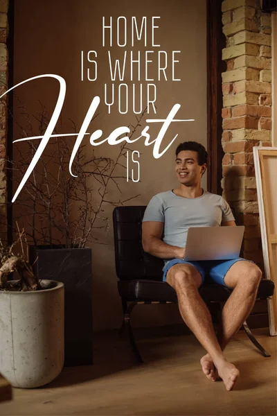 Happy mixed race freelancer chilling with laptop near lettering home is where your heart is lettering — Stock Photo