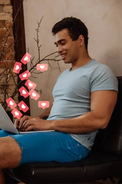 Sonriente freelancer de raza mixta utilizando un ordenador portátil cerca de corazones virtuales como gustos - foto de stock