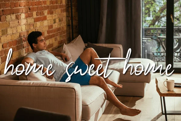 Bello uomo razza mista seduto sul divano in soggiorno vicino a casa dolce casa lettering — Foto stock