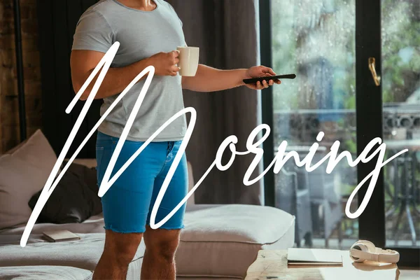 Cropped view of man with cup of coffee holding remote controller near morning lettering — Stock Photo
