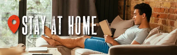 Encabezado del sitio web de hombre de raza mixta sonriente leer libro en el sofá cerca de quedarse en casa lettering - foto de stock