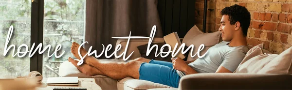 Website header of smiling mixed race man reading book on sofa near home sweet home lettering — Stock Photo