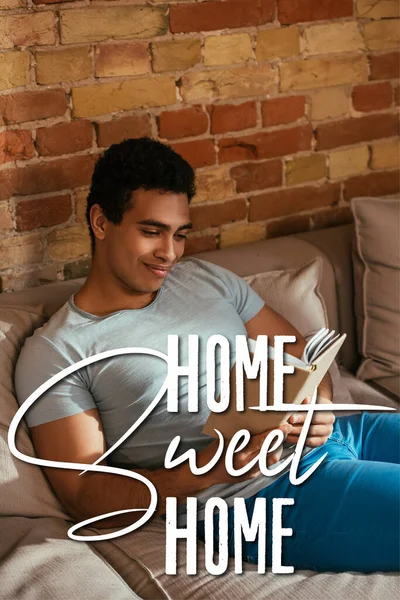 Happy mixed race man reading book on sofa near home sweet home lettering — Stock Photo