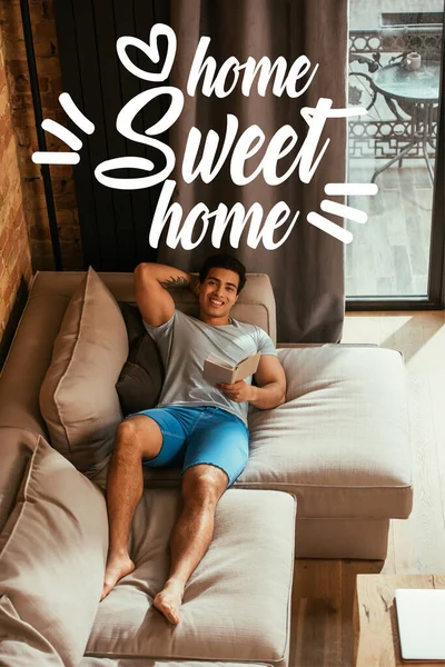 Happy mixed race man reading book while chilling on sofa near home sweet home lettering — Stock Photo
