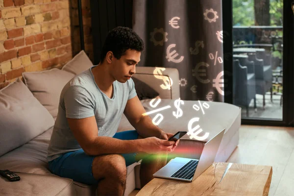 Handsome mixed race freelancer using smartphone near laptop and virtual money illustration — Stock Photo