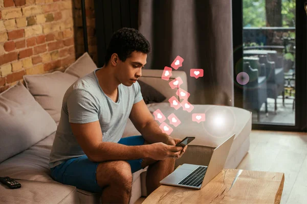 Handsome mixed race freelancer using smartphone near laptop and hearts as likes illustration — Stock Photo