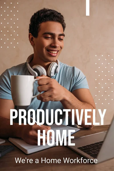 Hombre de raza mixta feliz sosteniendo la taza de café mientras escribía cerca de la computadora portátil y productivamente en casa, eran una escritura de mano de obra doméstica - foto de stock